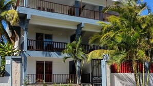 A multi-story building with balconies, a gated entrance, and surrounding palm trees. - Villa Devika 4, Rent Holidays Apartment (Trou aux Biches)