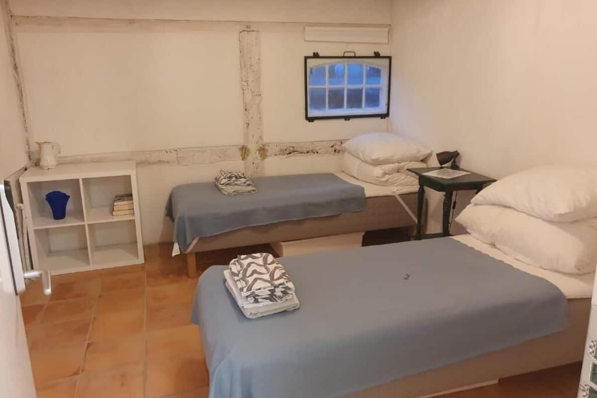 Quarto pequeno e simples com duas camas, uma mesinha, uma prateleira com um balde azul e uma janela com vista.