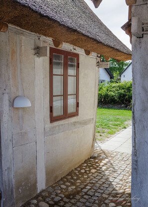 A building with a thatched roof, a window with wooden shutters, and a cobblestone pathway leading to a grassy area.