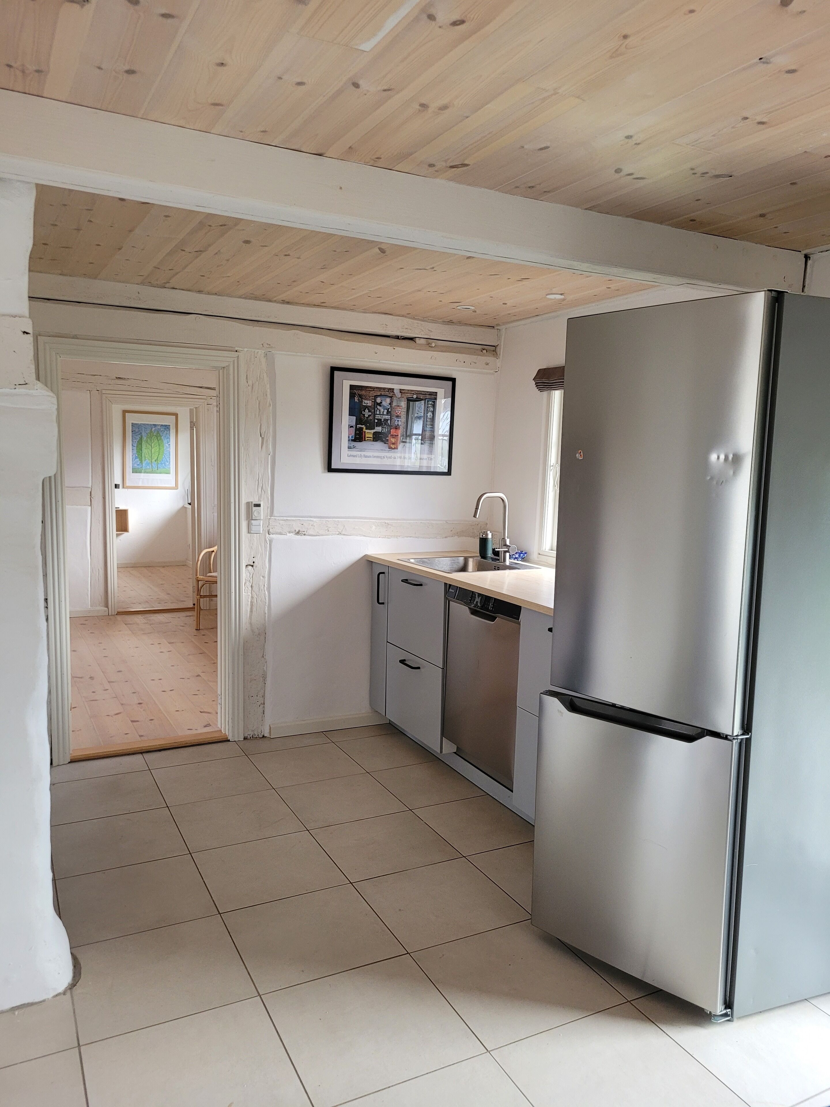 A compact kitchen with a stainless steel refrigerator, a sink, and a dishwasher. There is a framed picture on the wall and a wooden ceiling.