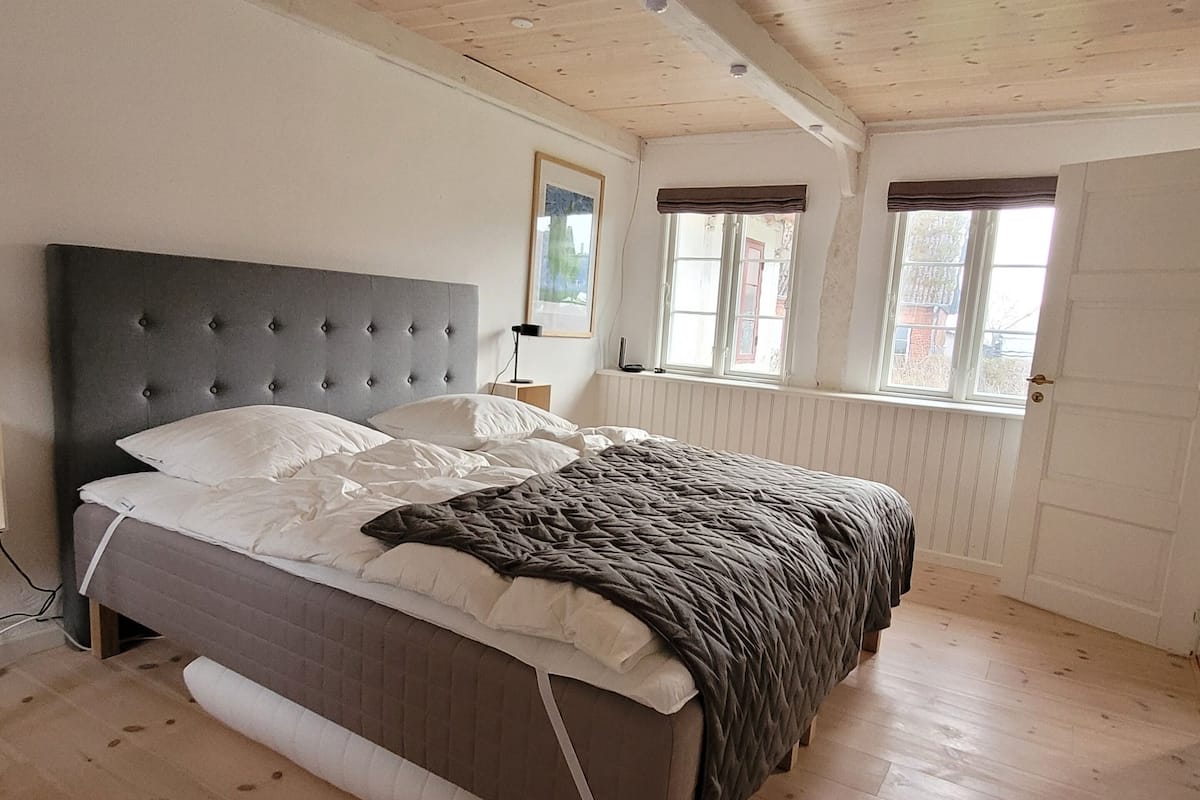 Quarto com cama, piso de madeira, paredes brancas e janela com vista.