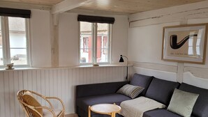 Una sala de estar acogedora con un sofá gris oscuro, un sillón de mimbre y una pequeña mesa de madera. Se ve una ventana con vistas a un edificio rojo.