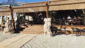 A beachside restaurant with outdoor seating, a stone facade, and a mountainous backdrop. - Olea Residence, Luxury sea View Apartment With Private Beach (Himarë)