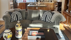 A living room with a grey sofa, a coffee table with decorative items, and a kitchen in the background. - Beautiful Home Walking Distance St Helena and Meadowood resort privileges. (St Helena)