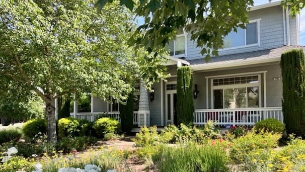 A two-story house with a front porch, surrounded by a garden with various plants and flowers. - Beautiful Home Walking Distance St Helena and Meadowood resort privileges. (St Helena)