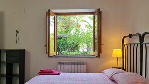 A bedroom with a bed, a window with an open view of greenery, a bedside lamp, and a radiator. - Nut House Apartment Near the sea in a Villa With Garden (Altavilla Milicia)