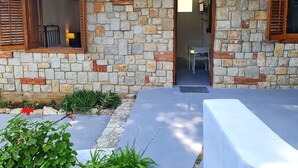 A stone building with a doorway leading to an interior room, a small outdoor seating area with a table and chairs, and a garden with plants and flowers. - Nut House Apartment Near the sea in a Villa With Garden (Altavilla Milicia)