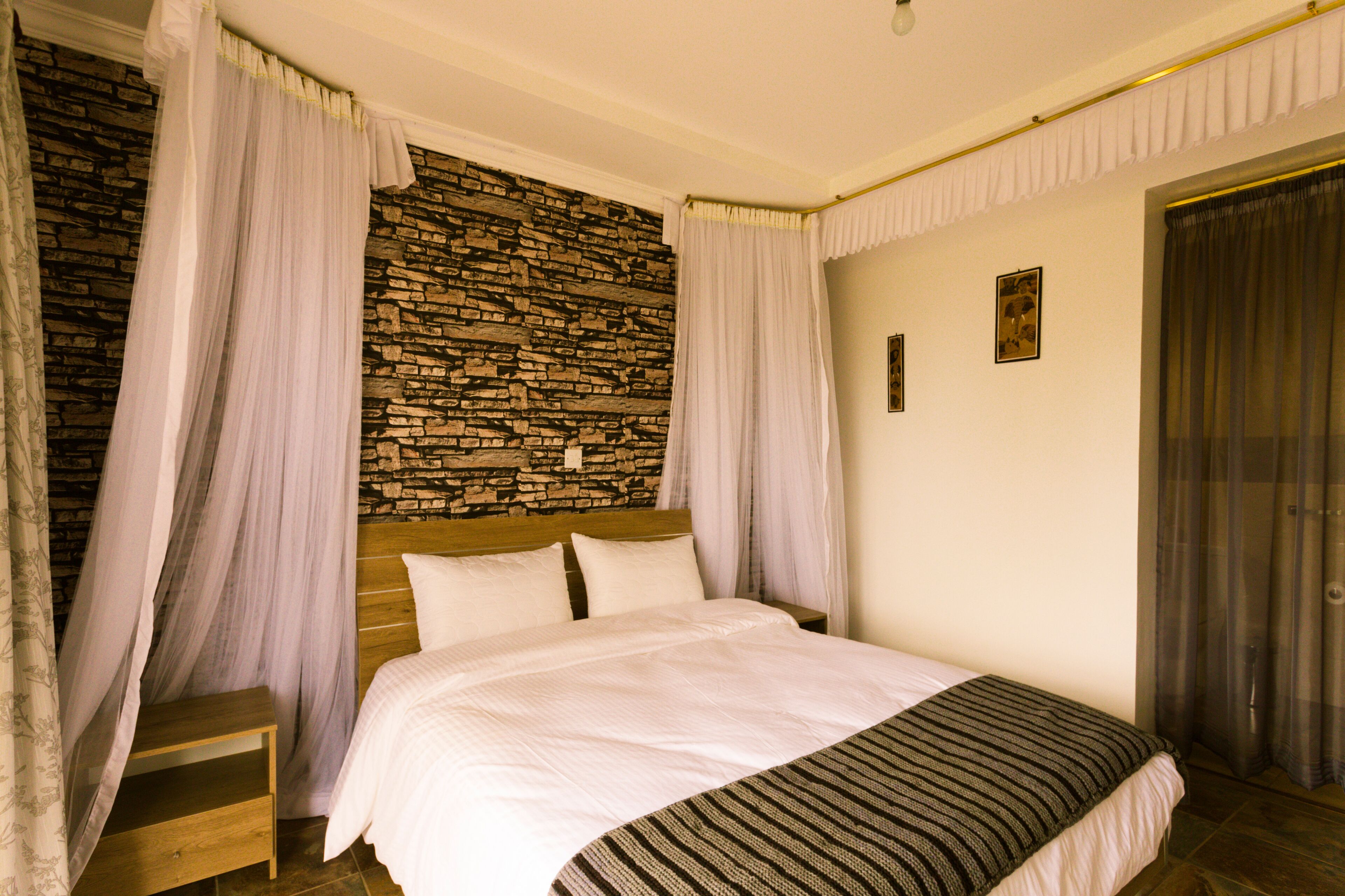 A bedroom with a stone wall, a wooden headboard, a bed with white bedding, a wooden nightstand, and sheer curtains.