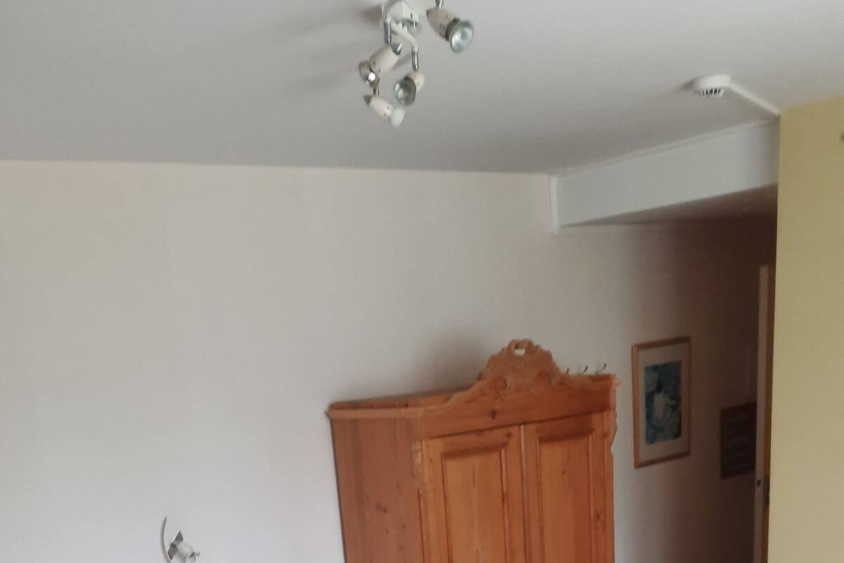 Quarto com cama de madeira, guarda-roupa de madeira, pequena mesa de madeira e cadeira de vime.
