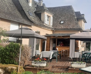 An outdoor patio area with wooden tables and chairs, a bar area, and stone planters with flowers.