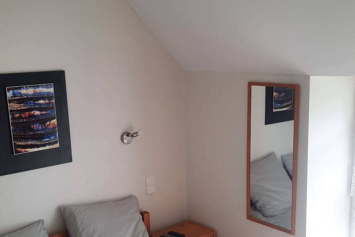 Quarto com cama, mesa de cabeceira, espelho fixado na parede e luminária no teto.