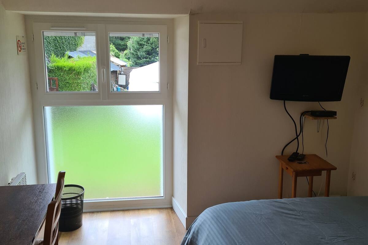 Quarto com cama, mesa com cadeiras, televisão em um suporte e janela com vista para o verde.