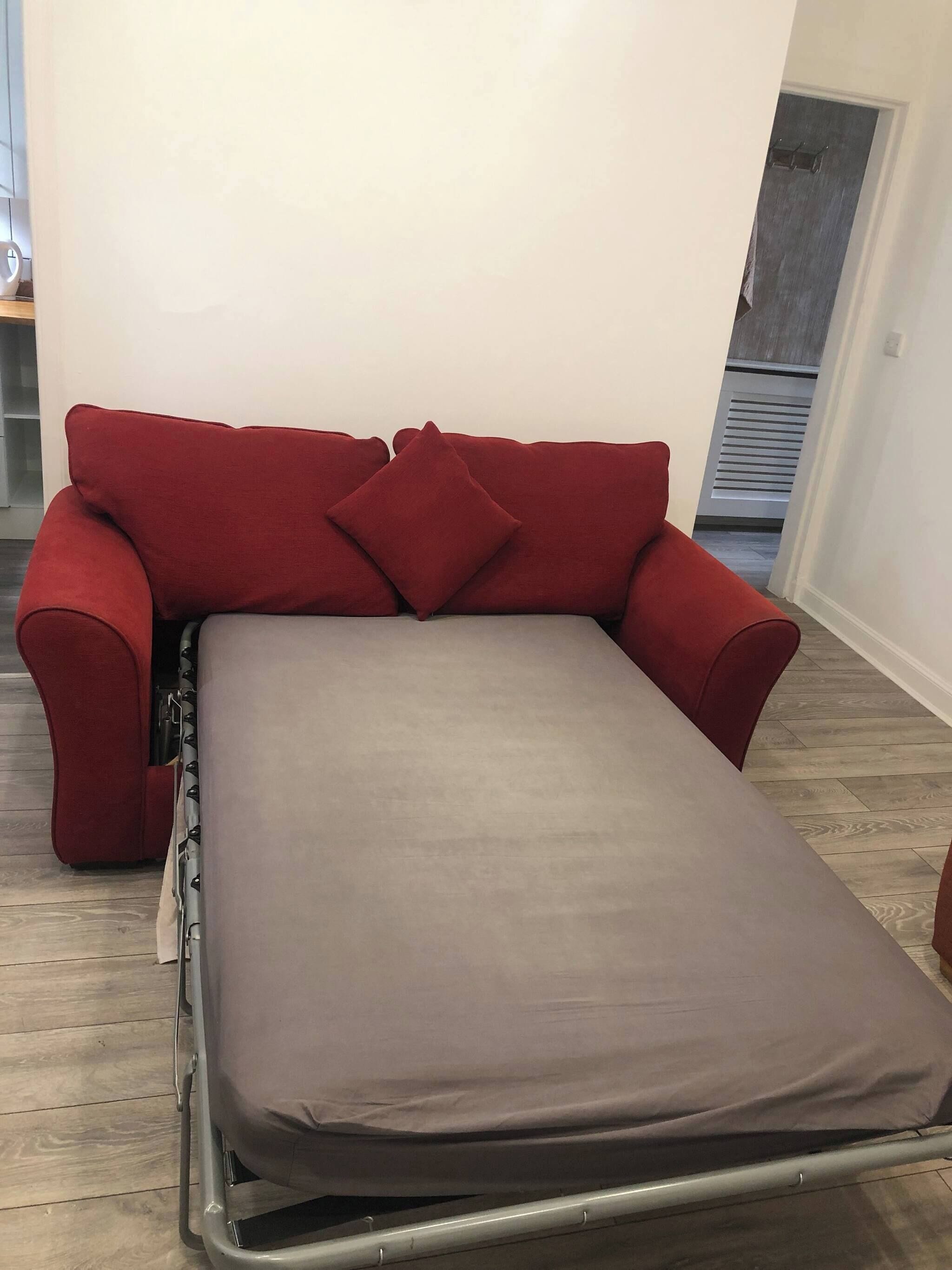 Un sofá rojo con un colchón gris, una mesita pequeña y una pared blanca.