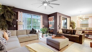 Ein Wohnzimmer mit beiger Couch, braunem Ledersofa und einem Couchtisch. Es gibt einen Deckenventilator, ein Fenster mit Vorhängen und eine Pflanze.