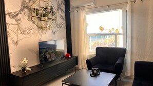 Ein modernes Wohnzimmer mit einem großen Flachbildfernseher, einem dunklen Holzkonsole, einem schwarzen Sessel und einem Couchtisch. Auf der Konsole steht ein dekoratives sechseckiges Regal mit Pflanzen und einer roten Vase. Durch ein Fenster ist ein Blick auf Palmen und einen Strand möglich.