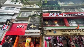 Vista da rua com vários edifícios comerciais, incluindo um restaurante KFC, um hotel chamado 'Olive' pela Taj Hotels & Resorts, e uma loja chamada 'Tea Junction'.