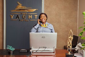 A reception desk at The Yacht Hotel with a computer, a desk lamp, a plant, and a decorative award. - The Yacht Hotel (Lekki)