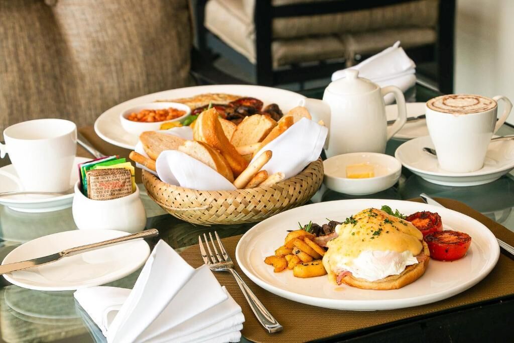 A breakfast setting with a plate of eggs benedict, toast, and roasted tomatoes, a coffee cup, and a basket of bread.