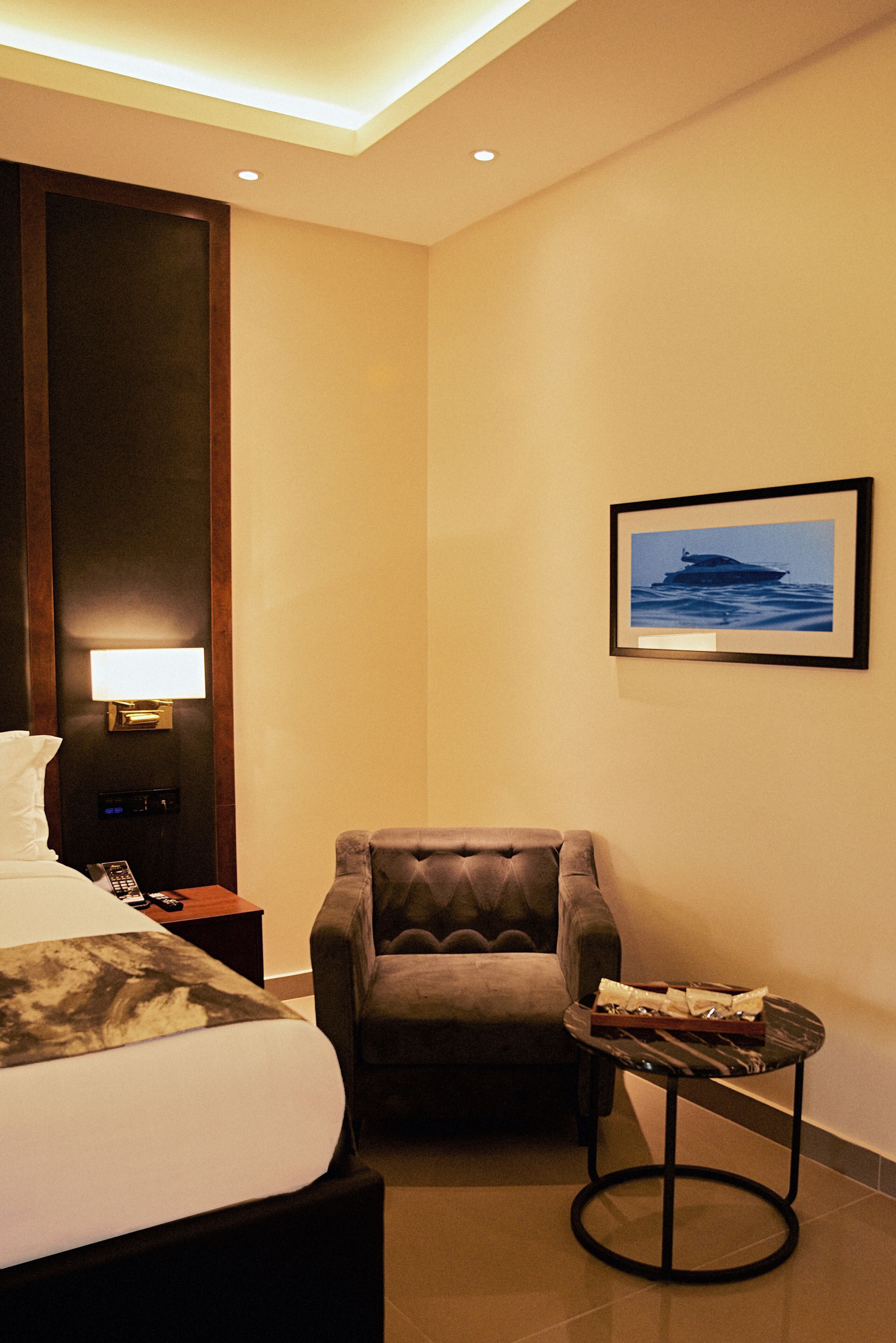 A hotel room with a bed, a chair, a small table, a wall-mounted lamp, a framed picture, and a nightstand with a phone.