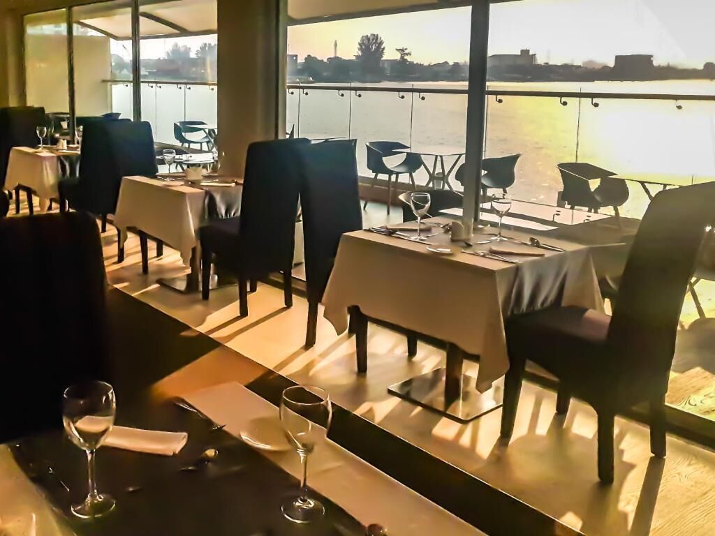 A restaurant with tables set for dining, a view of a body of water, and a cityscape in the background.