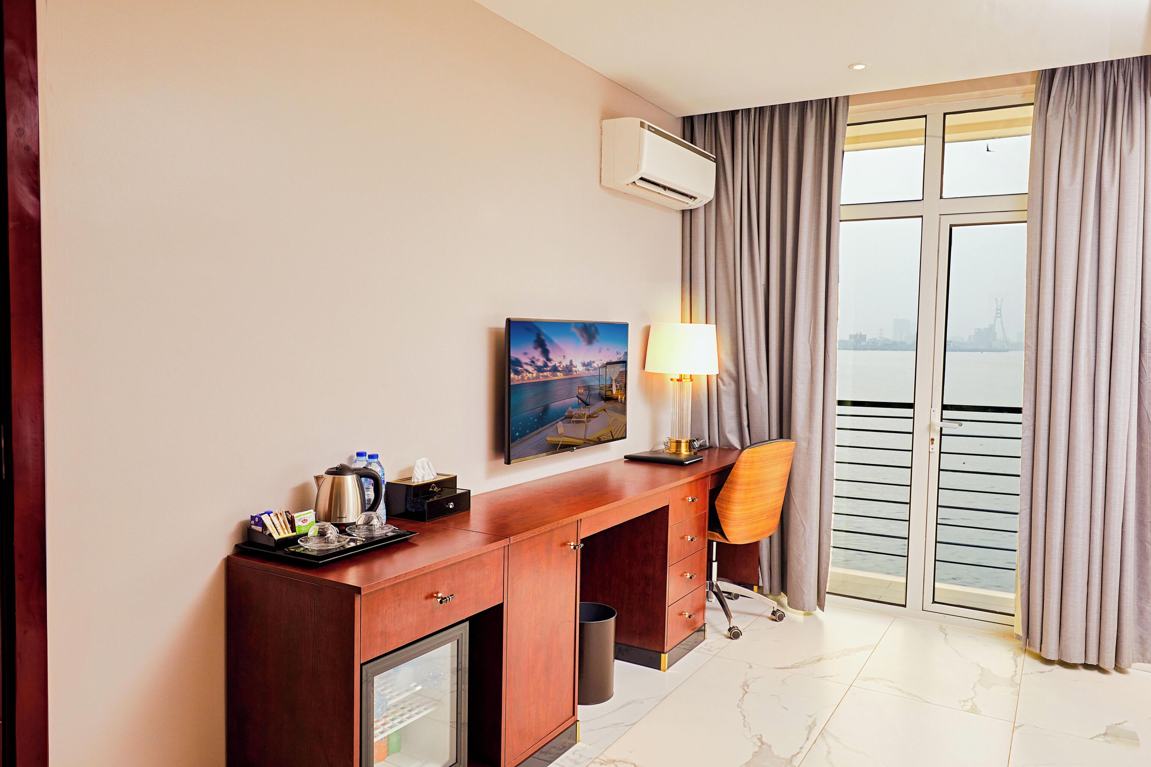 Un cuarto de hotel con un escritorio de madera, una lámpara, un cuadro en la pared y vistas al agua por la ventana.