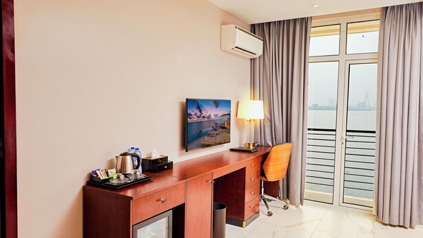 A hotel room with a wooden desk, a lamp, a picture on the wall, and a view of the water through the window. - The Yacht Hotel (Lekki)