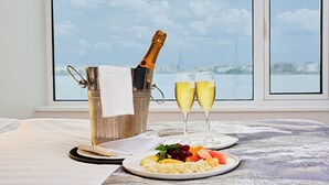 A champagne bottle in a metal bucket, two glasses of champagne, and a plate of fruit on a table by a window with a view of a body of water and a bridge. - The Yacht Hotel (Lekki)