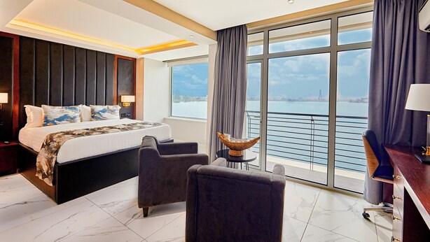 A modern hotel room with a large bed, two armchairs, a desk, and a view of the sea. - The Yacht Hotel (Lekki)