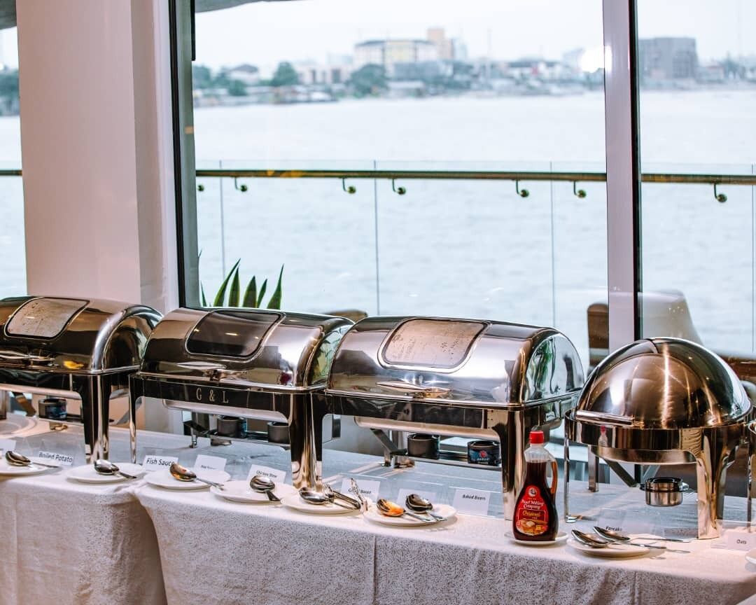 A buffet setup with various food items and sauces, a view of a body of water through large windows, and a cityscape in the background.