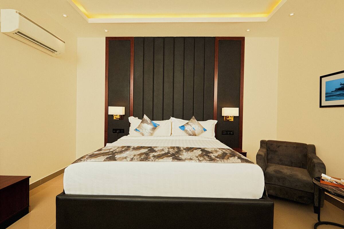 A hotel room with a large bed, two bedside lamps, a chair, and an air conditioning unit.