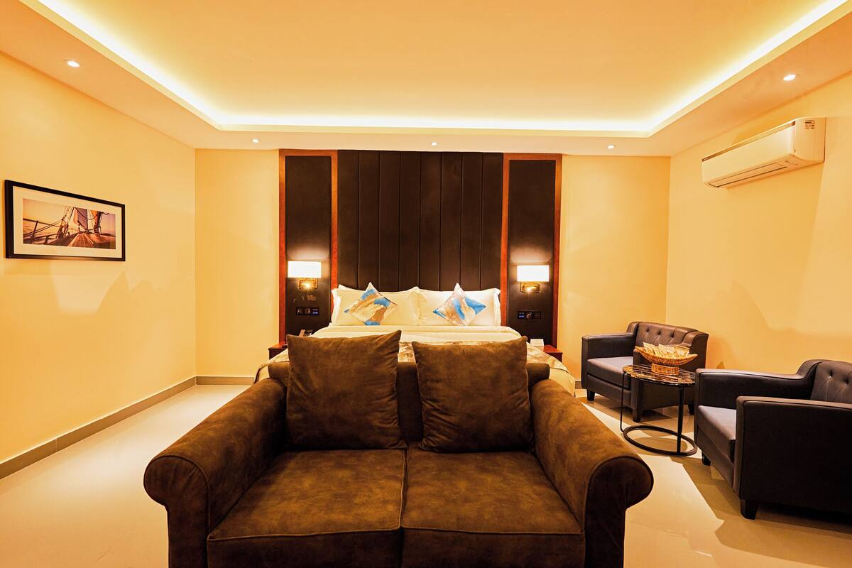 A hotel room with a bed, two sofas, a small table, and a wall-mounted air conditioner.