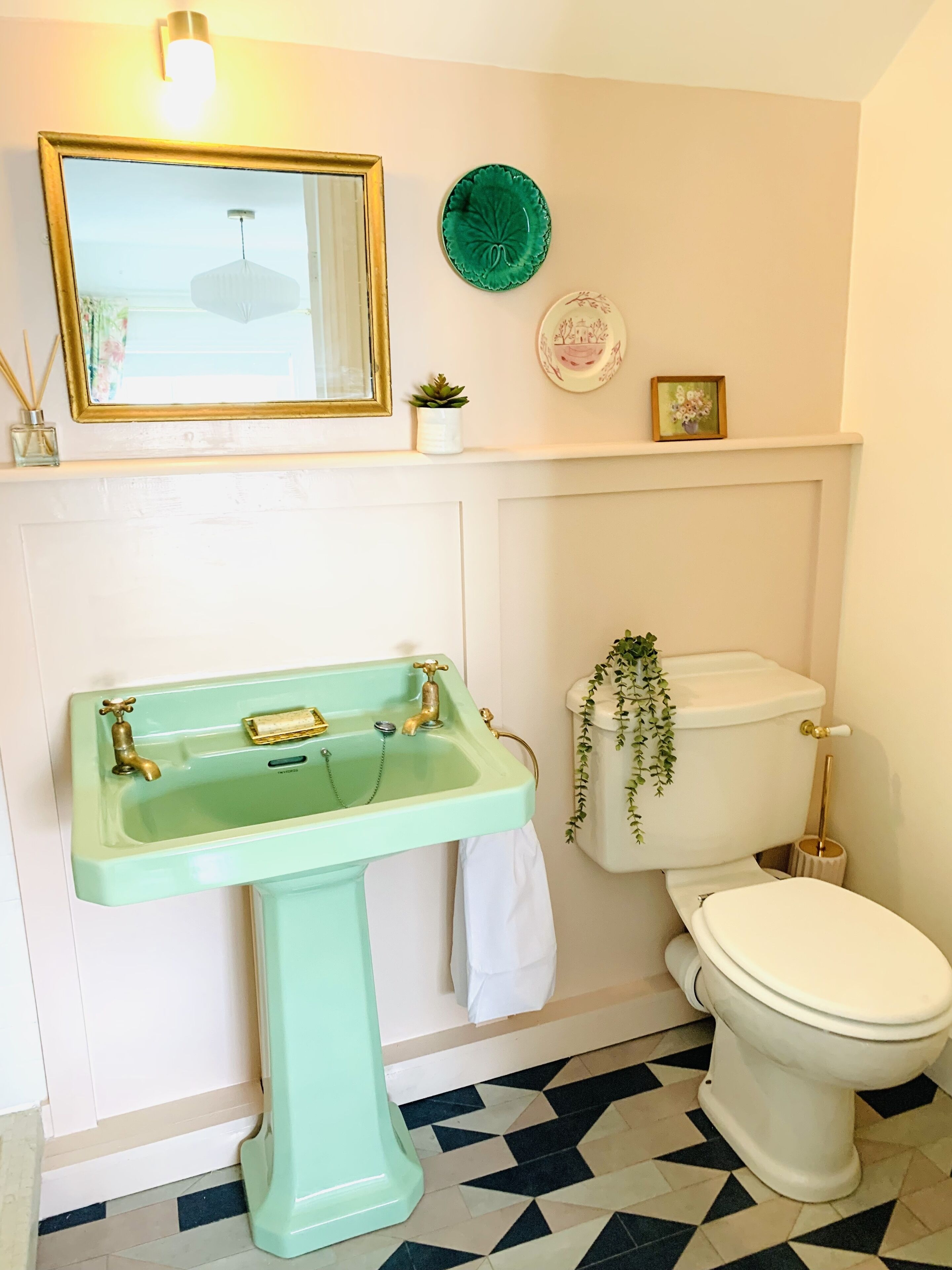A bathroom with a green pedestal sink, a white toilet, a mirror with a gold frame, and a green decorative plate on the wall.
