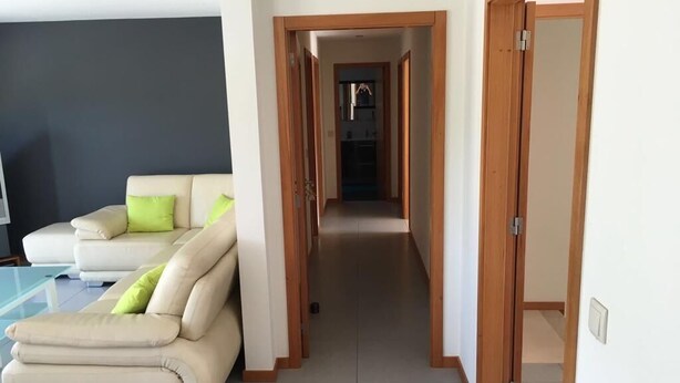 A modern living room with a beige sofa and green cushions, a glass coffee table, and a wooden door leading to a hallway. - Bright, modern house with green views! (Lage)