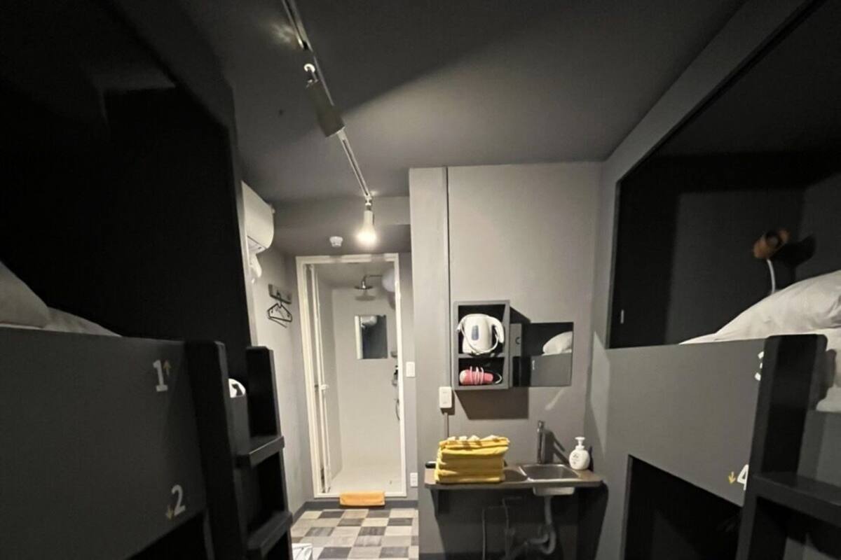 A dormitory room with bunk beds, a desk, a chair, a small mirror, and a shelf with toiletries.
