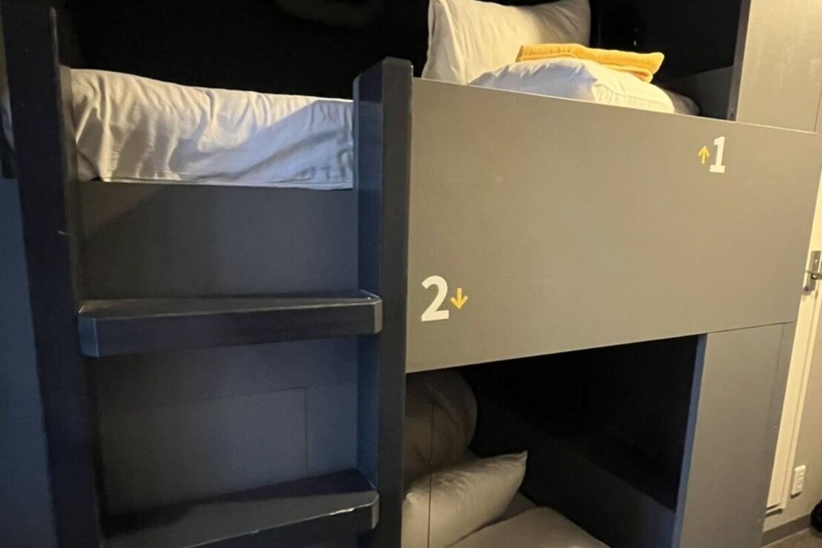A bunk bed with two levels, each with a pillow and a blanket. There is a shelf unit attached to the left side of the bed.