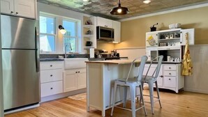 Una cocina con armarios blancos, un refrigerador de acero inoxidable, piso de madera y una lámpara colgante.