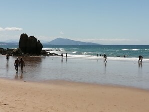 Een strand met mensen die wandelen en in het water spelen, een grote rotsformatie en een berg op de achtergrond.
