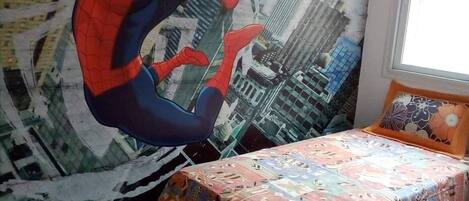 Un dormitorio con un mural de Spider-Man en la pared, una cama con colcha colorida y una ventana con vistas a edificios.