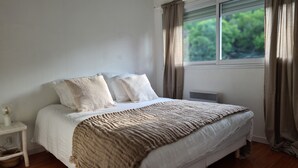 Ein Schlafzimmer mit einem Bett, weißer Bettwäsche, einem Nachttisch, einer Lampe und einem Fenster mit Vorhängen.