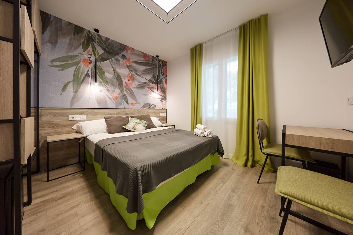 A modern hotel room with a large bed, a desk, and a chair. There is a painting on the wall and a window with curtains.