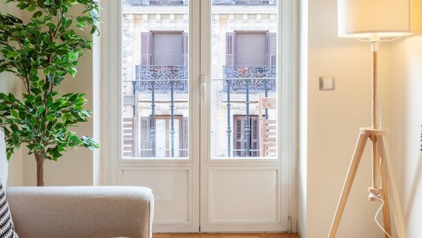 Quarto com porta branca, planta em vaso, luminária de chão e vista de edifícios pela porta.