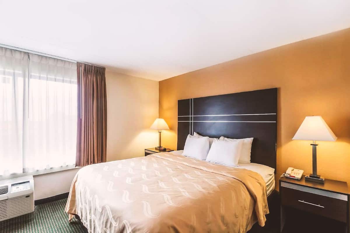 A hotel room with a large bed, two bedside tables with lamps, a window with curtains, and a wall-mounted air conditioning unit.