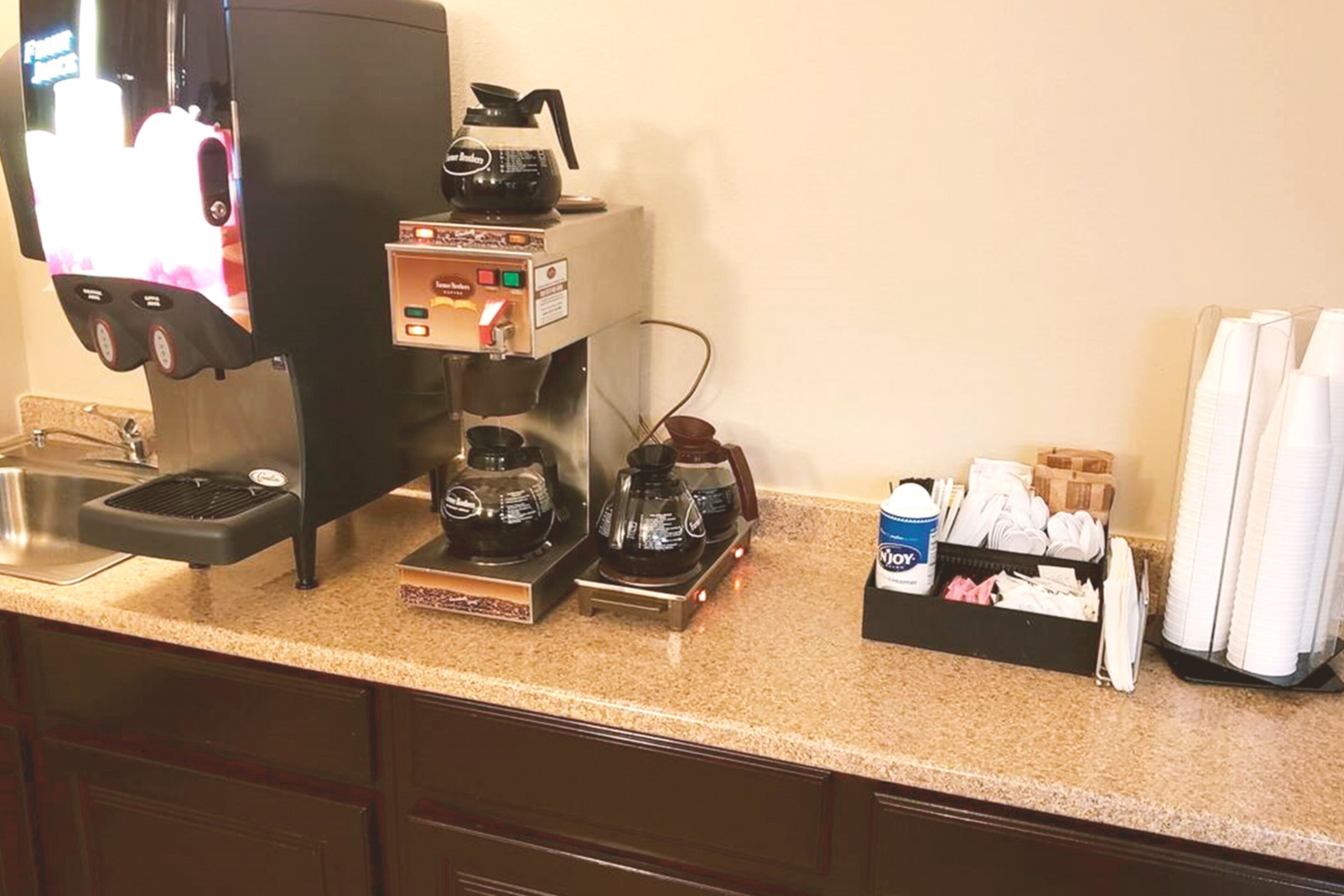Un coin café équipé d’une machine à café, d’un moulin et d’une cafetière. On y trouve des tasses et une boîte de dosettes de café sur le comptoir.
