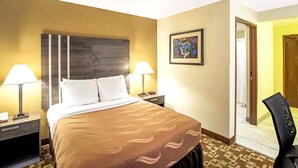 Ein Hotelzimmer mit Bett, zwei Nachttischlampen, einem Nachttisch, einem gerahmten Bild an der Wand und einer Tür zu einem weiteren Raum.