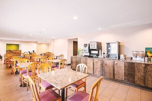 Una sala da pranzo di un hotel con diversi tavoli e sedie, un angolo caffè e un bancone con vari elettrodomestici.