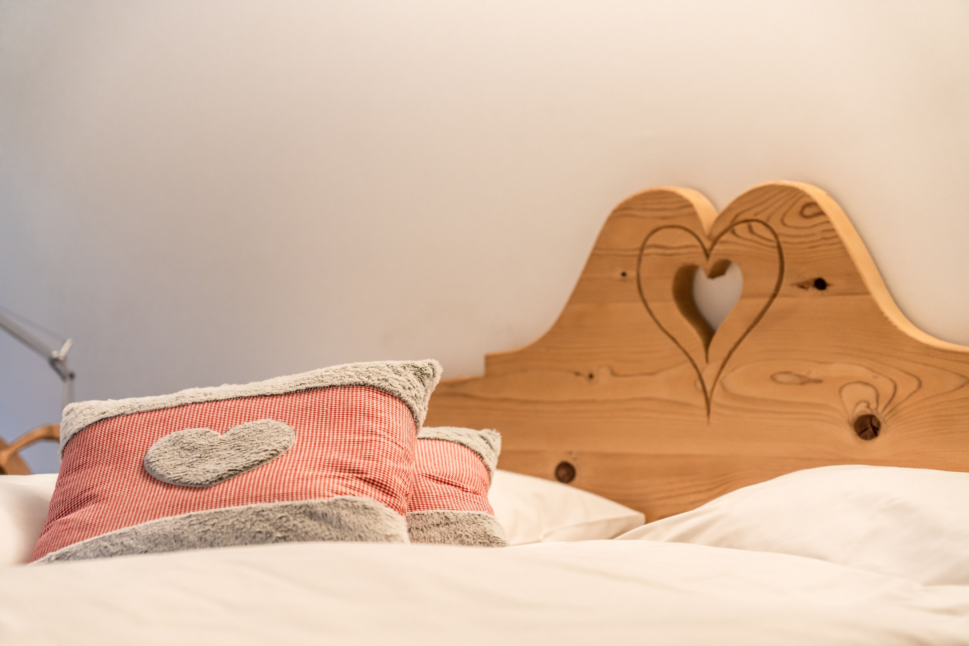 Un lit avec une tête de lit en bois ornée d’un motif en forme de cœur, un coussin à carreaux rouges et blancs, et un coussin moelleux de couleur grise.