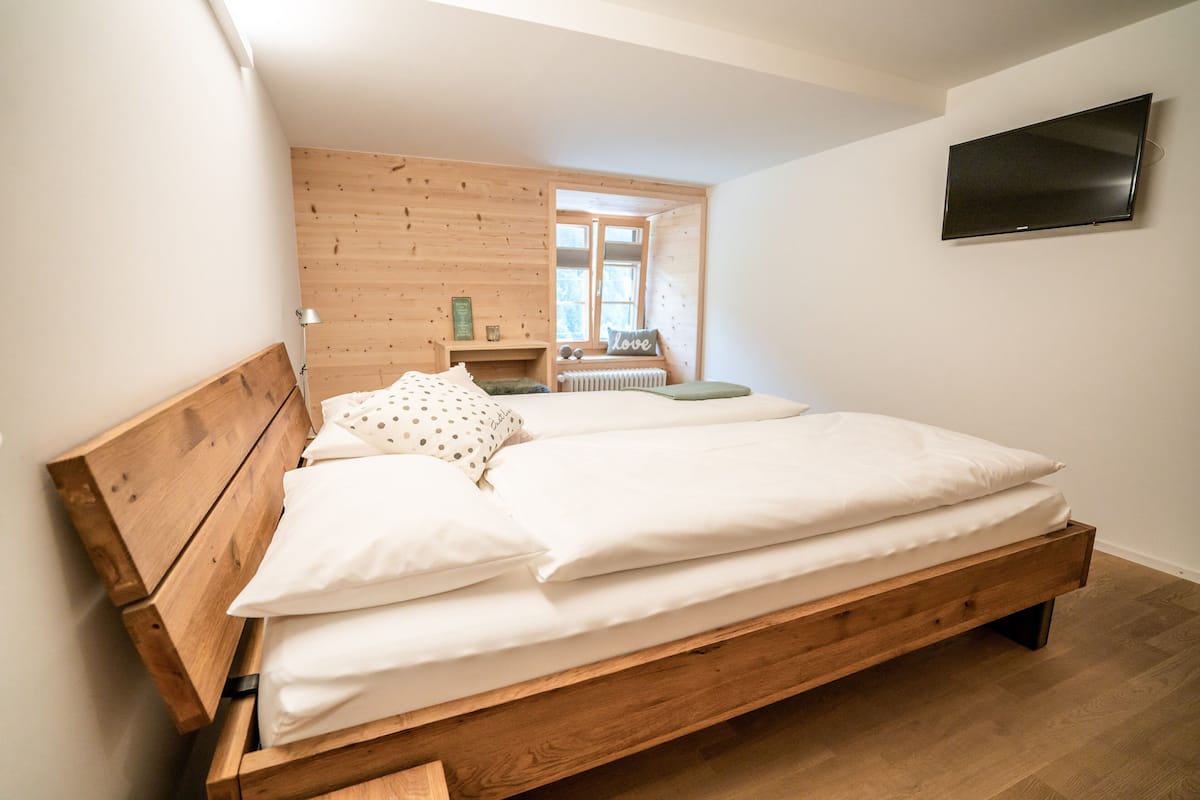 Une chambre à coucher avec un lit en bois, un téléviseur fixé au mur et une fenêtre donnant sur l’extérieur.