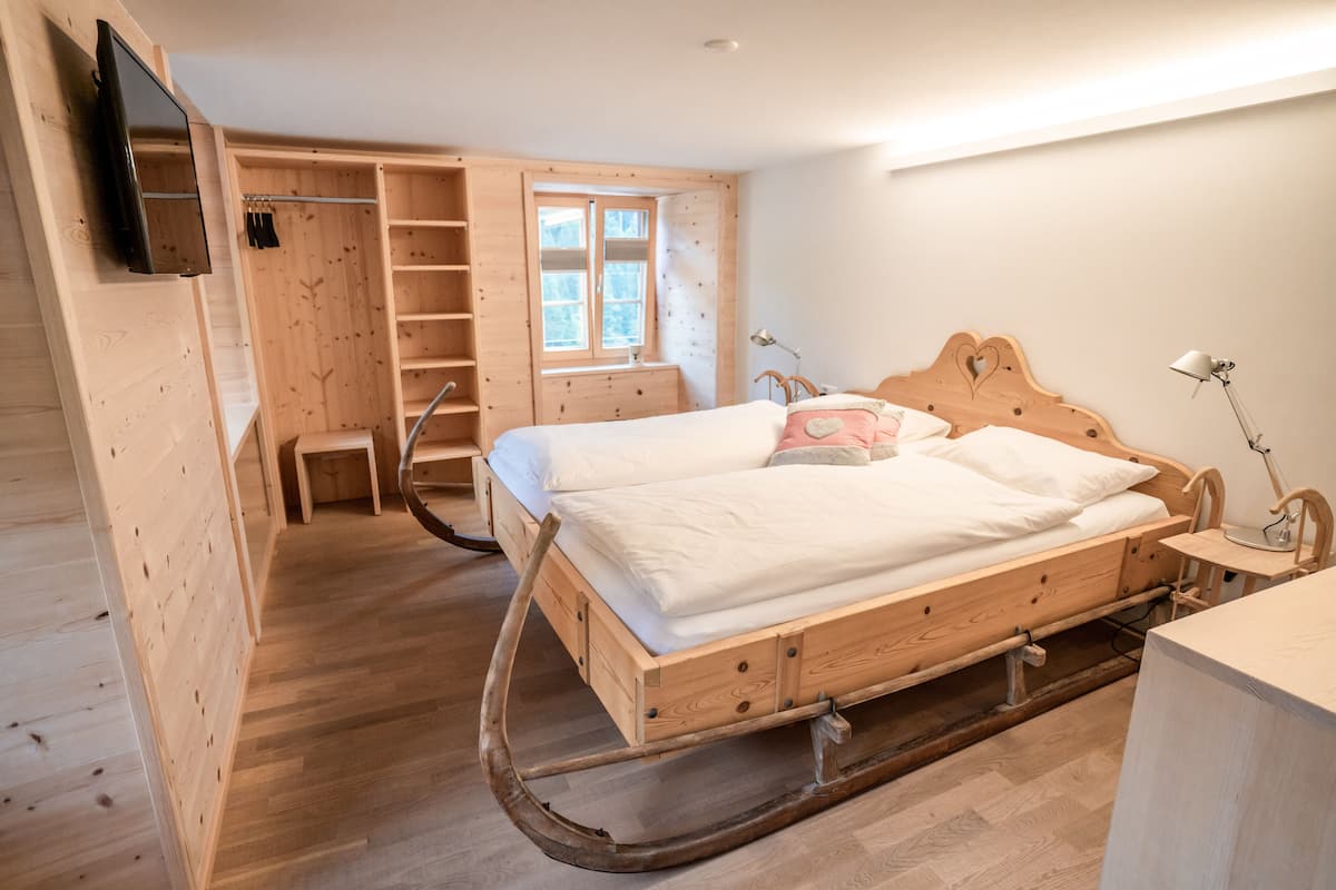 Un lit en bois avec un élastique, une armoire en bois, une télévision et une fenêtre donnant sur des arbres.