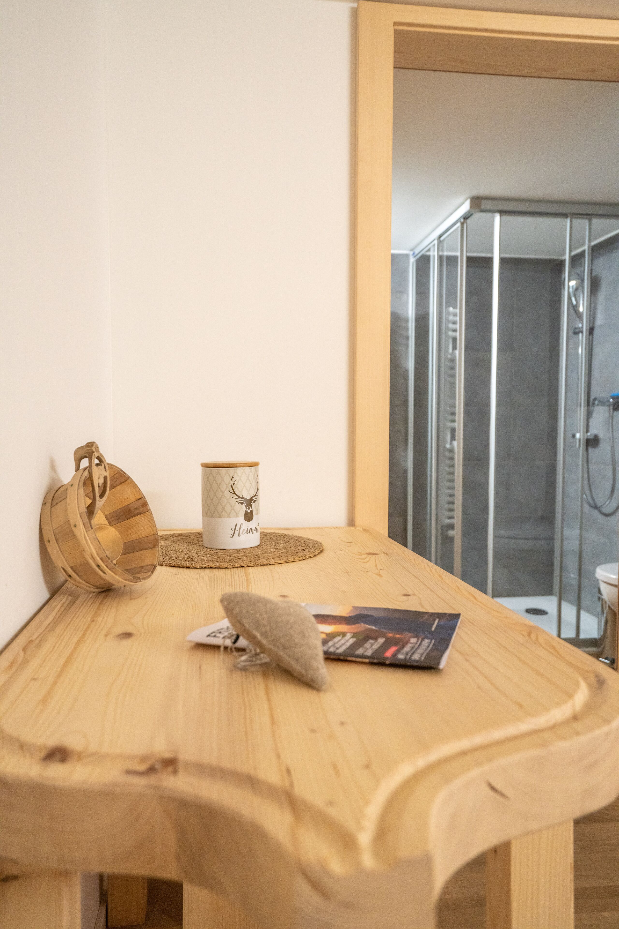 A wooden table with a woven placemat, a wooden bag, a mug with a deer design, and a magazine. A shower area is visible in the background.