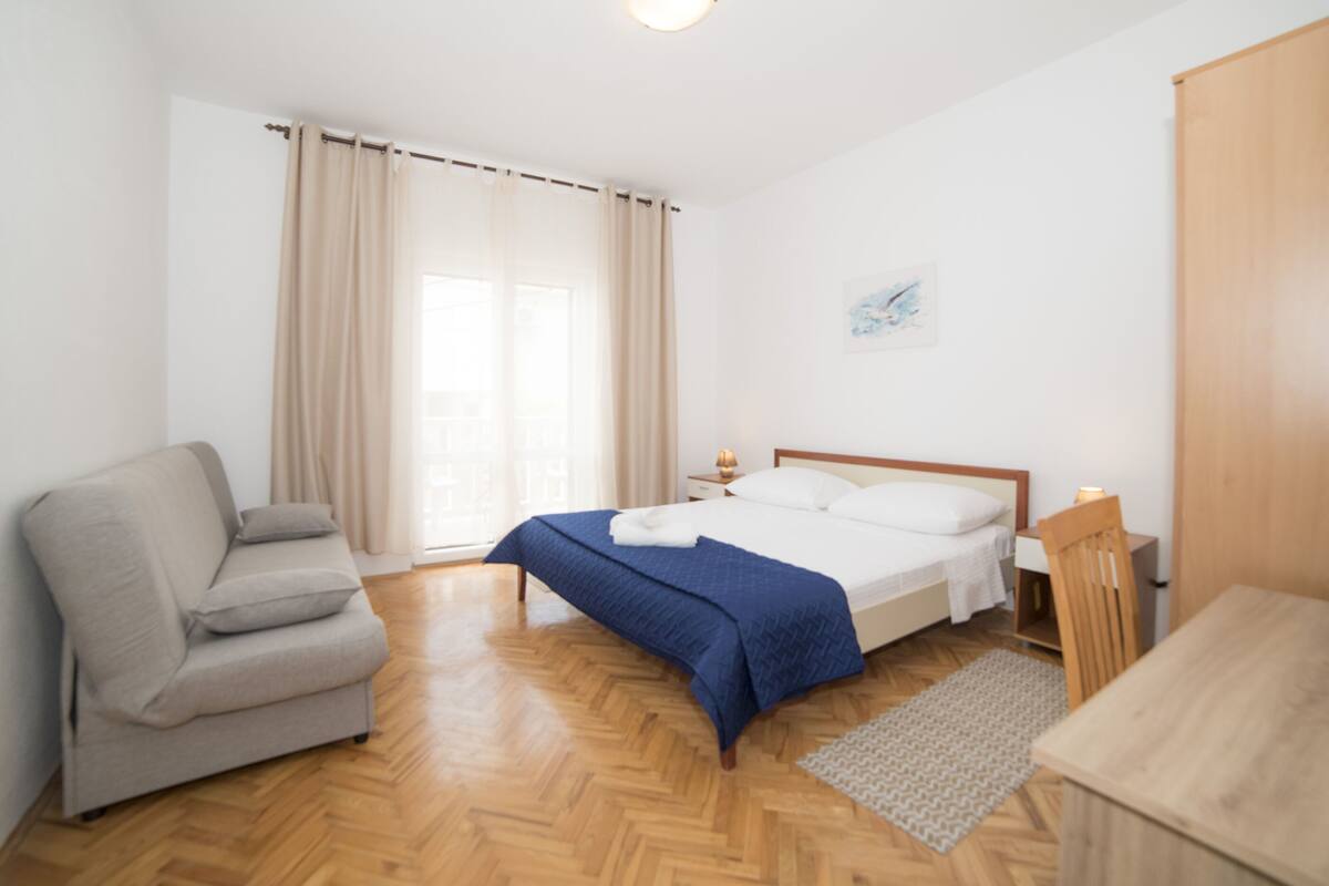 A bedroom with a bed, a sofa, a wooden table, and a window with curtains.